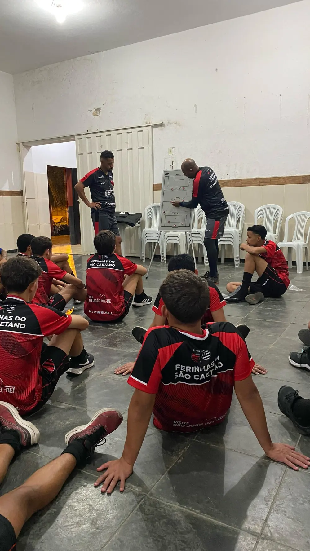 Un entrenador explica una jugada en una pizarra táctica a un grupo de chicos sentados en el piso