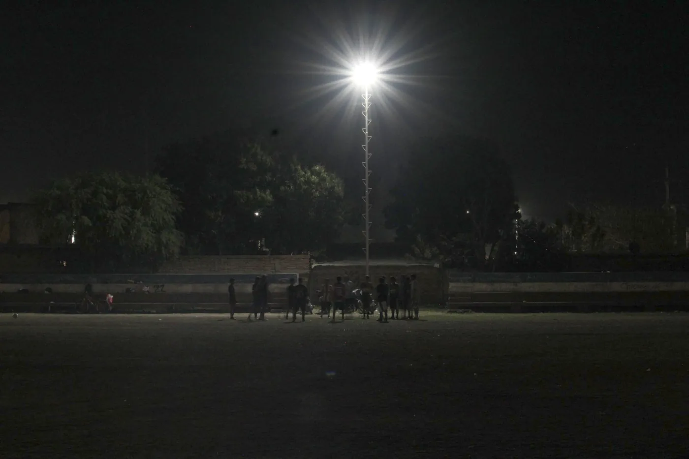 Una torre de iluminación encendida de noche sobre la cancha, con un grupo de chicos debajo
